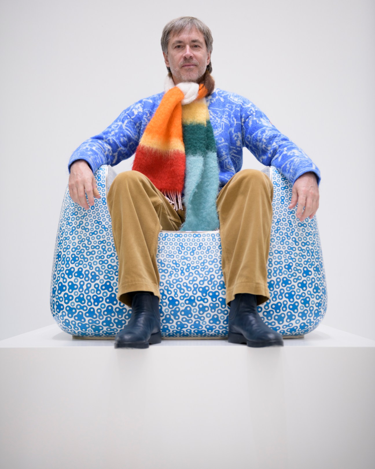 Man with beard sitting on blue patterned bean bag chair wearing colorful striped scarf and blue sweater