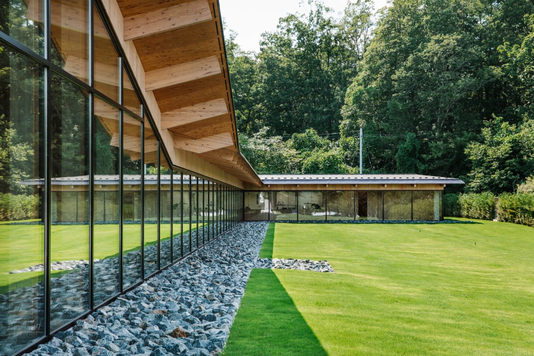 Modern glass-walled building with exposed wooden beam ceiling overlooking landscaped grounds with gravel beds and lawn