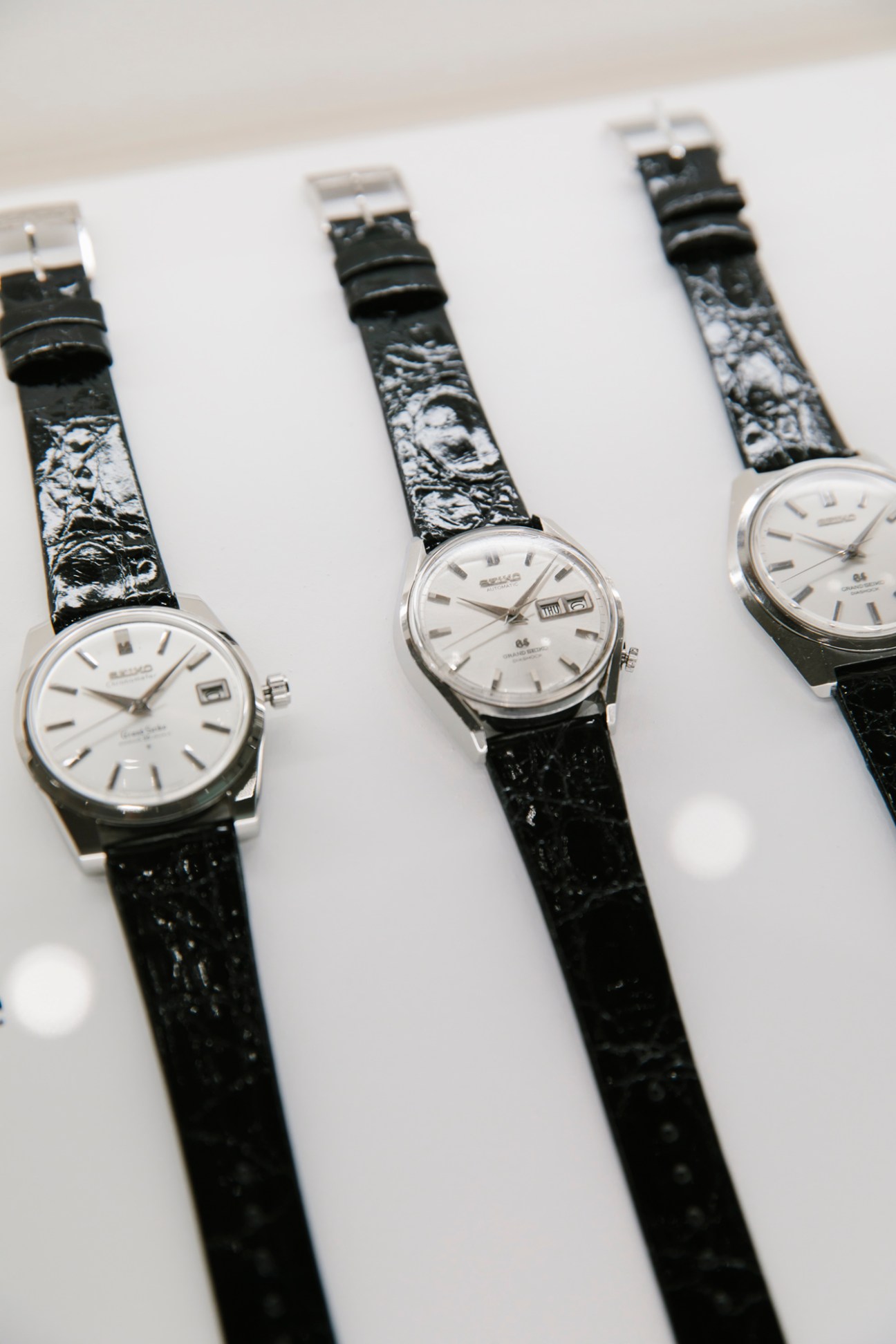 Three vintage Grand Seiko watches from 1964 and 1967 with silver faces and black leather straps displayed on white