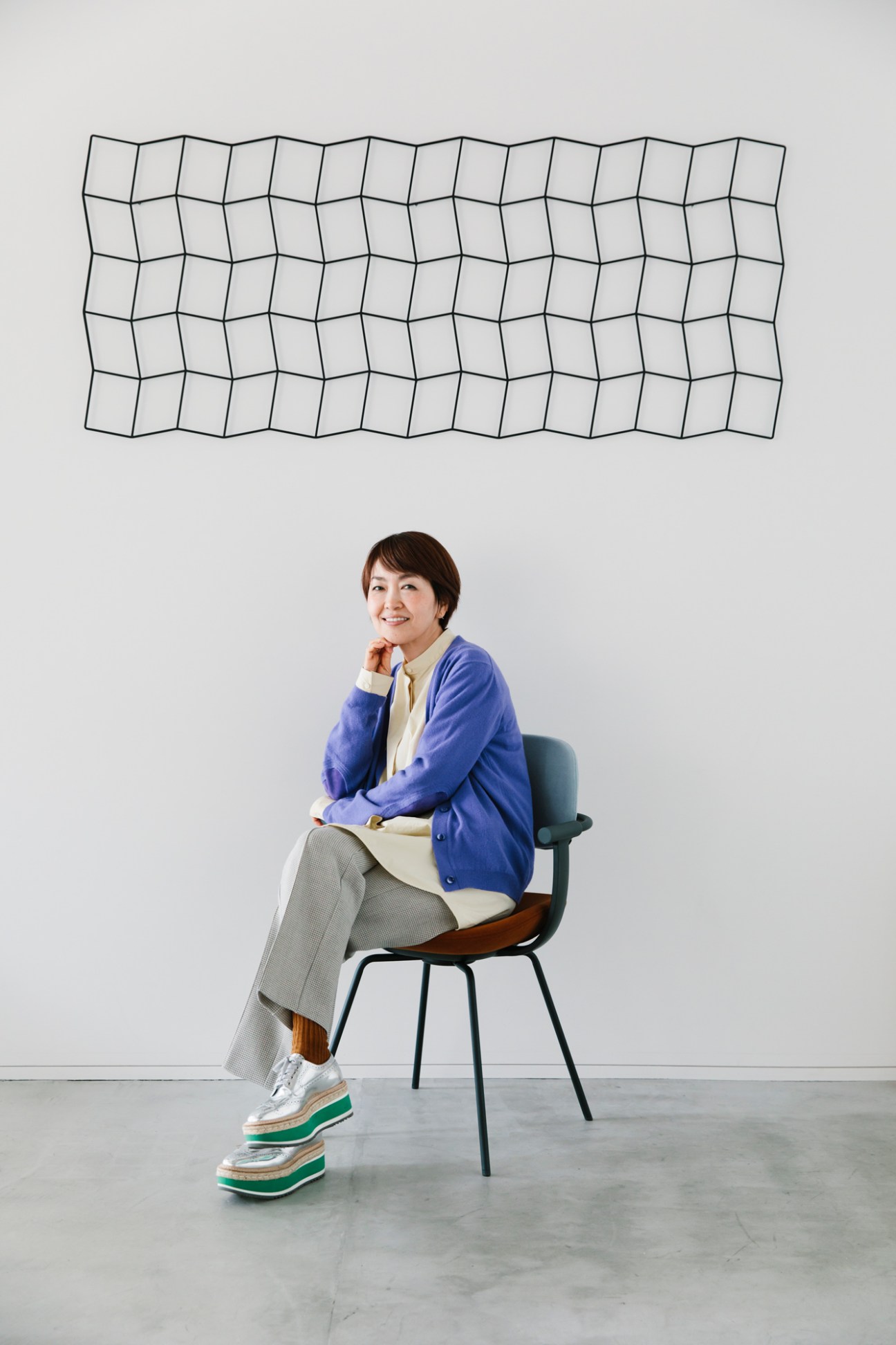 Fumie Shibata sitting in a modern chair wearing a blue cardigan and gray pants, with geometric wall art behind her