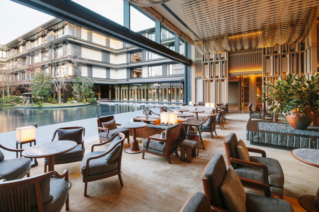 Sun-lit lobby lounge with wooden furniture, plants, and view of pool and modern hotel building