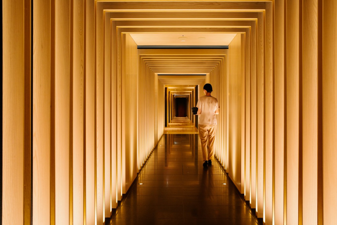 Person in white clothing walking through a warmly lit wooden corridor with repeated geometric architectural frames