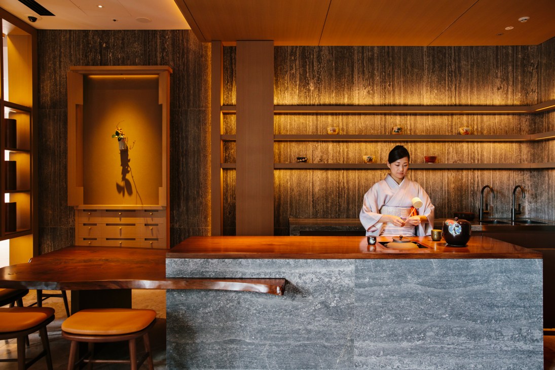 Woman in pink kimono performing tea ceremony at wooden counter in modern Japanese interior with textured walls