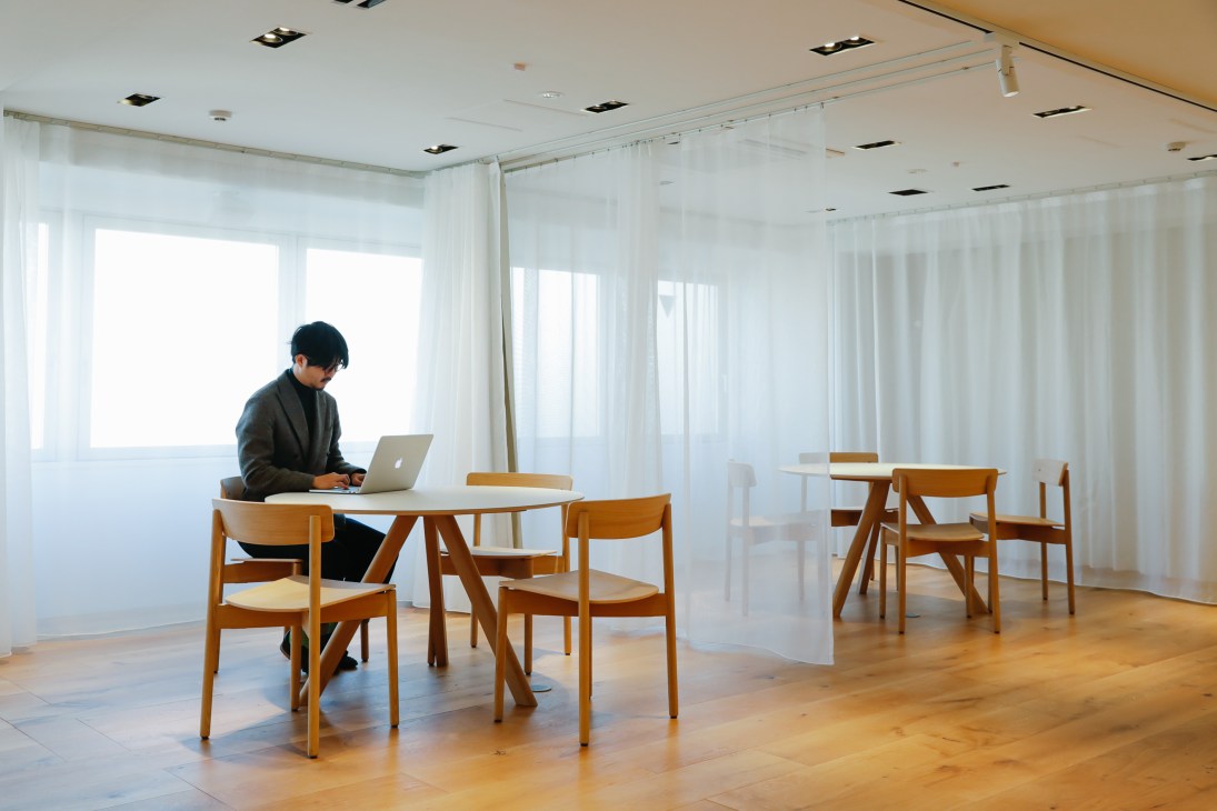 Person working on laptop at wooden table in minimalist white office space with natural lighting