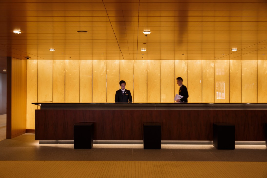 Two hotel staff members in dark suits working behind a long wooden reception desk in a modernist lobby