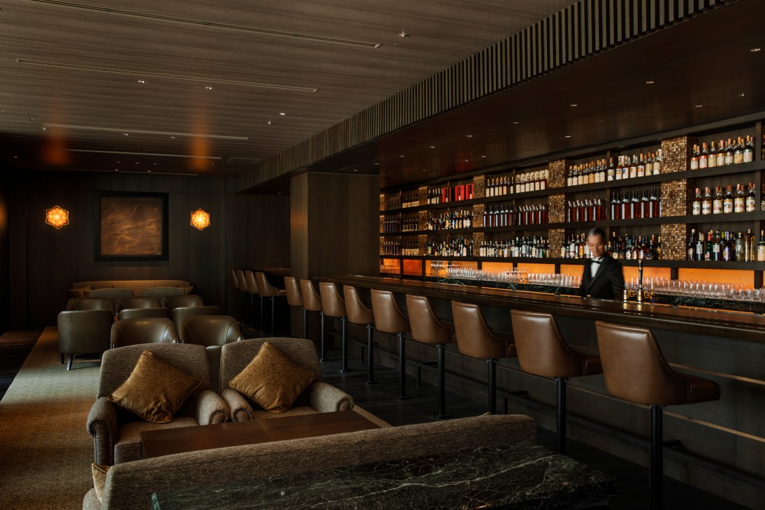 Elegant hotel bar with brown leather stools, extensive whiskey collection, and bartender in formal attire