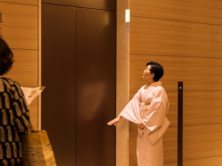 A woman in a pale pink kimono holds open an elevator door in an elegantly wood-panelled hotel lobby.