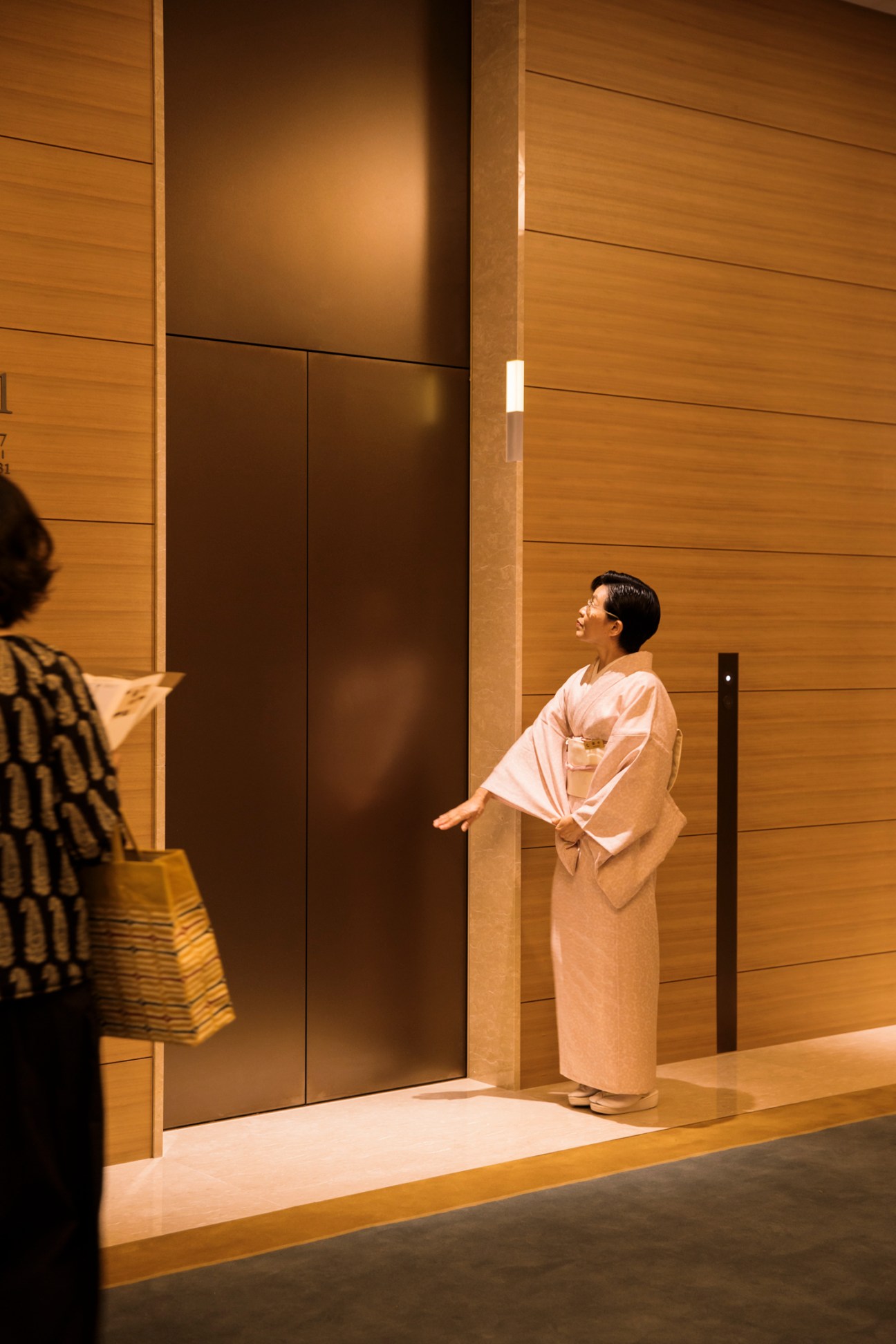 Woman in traditional kimono waiting by elevator in modern hotel lobby with warm wood paneling