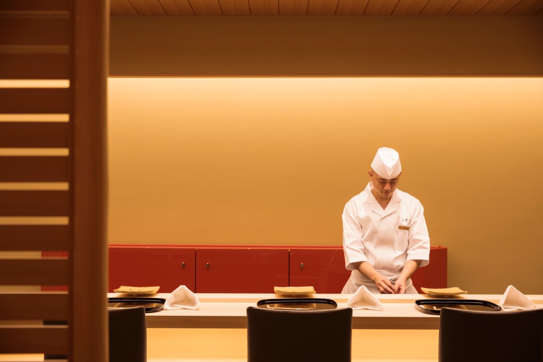 Japanese chef in white uniform and traditional hat preparing food at minimalist sushi counter in modern restaurant