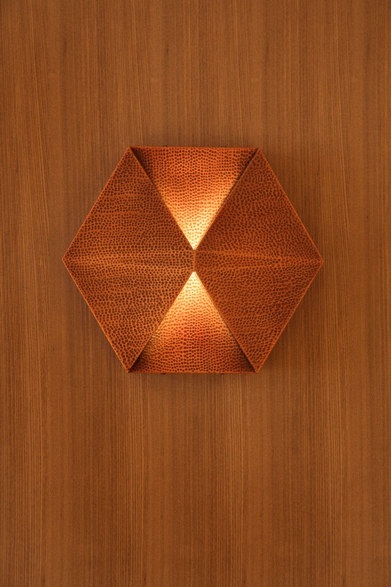 Hexagonal copper wall lamp with perforated pattern mounted on wood paneling, casting warm triangular light