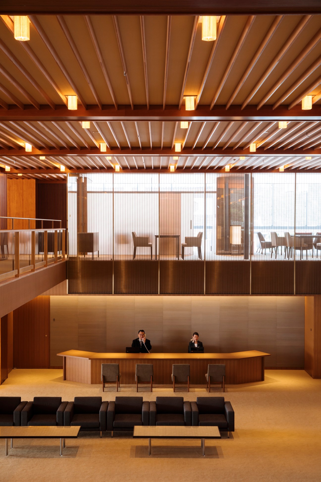 Modern hotel lobby with wood-paneled concierge desk, reception staff, and seating area under geometric ceiling