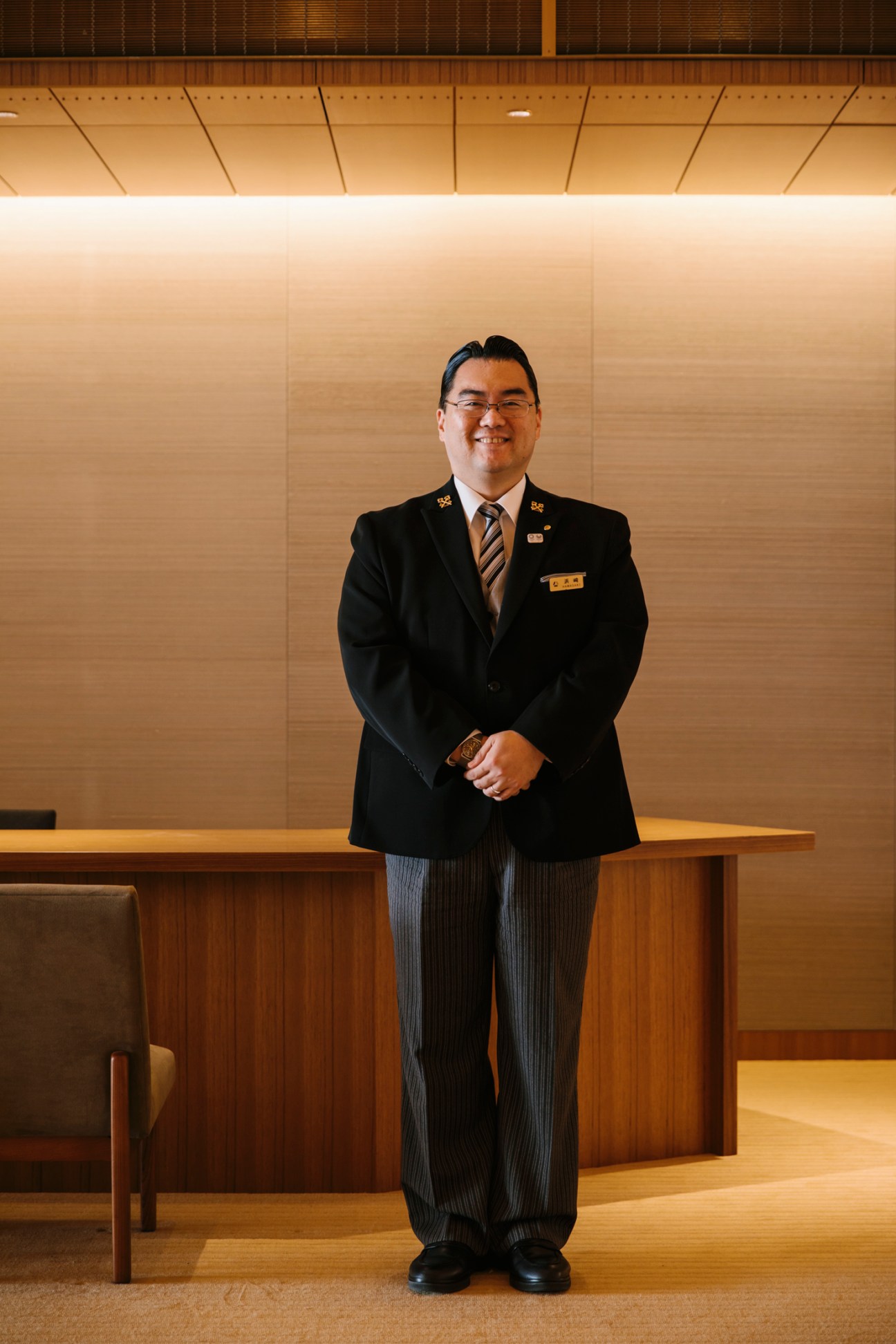 Smiling hotel staff member in formal uniform standing in modern wood-paneled lobby interior