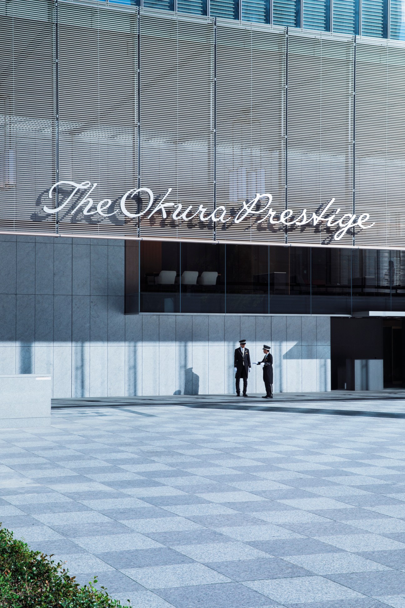 Two uniformed staff members stand outside the modern glass facade of Hotel Okura's Prestige Tower entrance