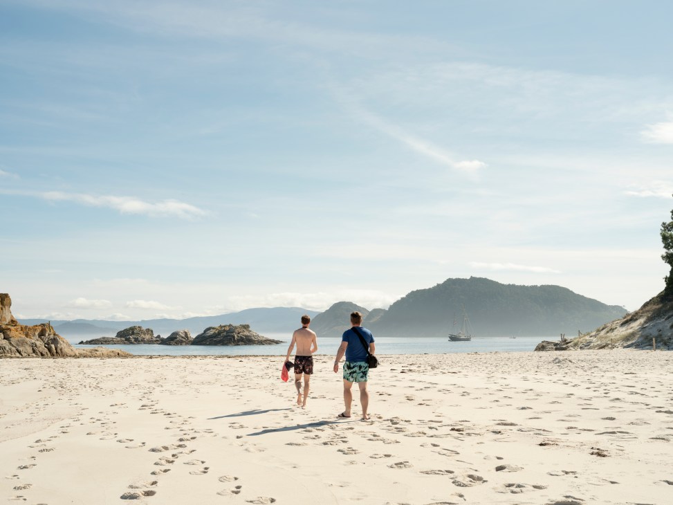 Two men walking on a sandy beach with rocky islands and mountains visible across the water.