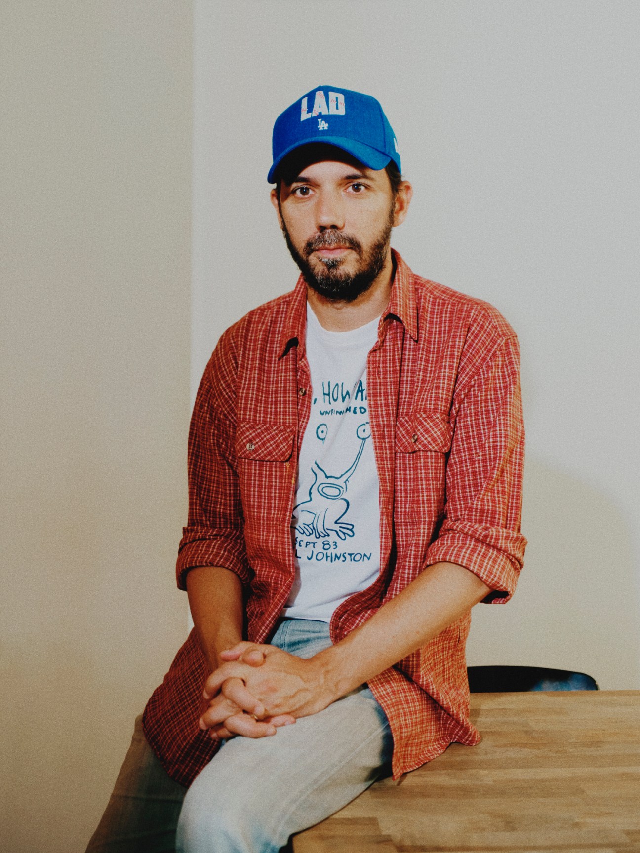 Franck Annese wearing a blue LA cap and red plaid shirt sitting casually on a wooden surface