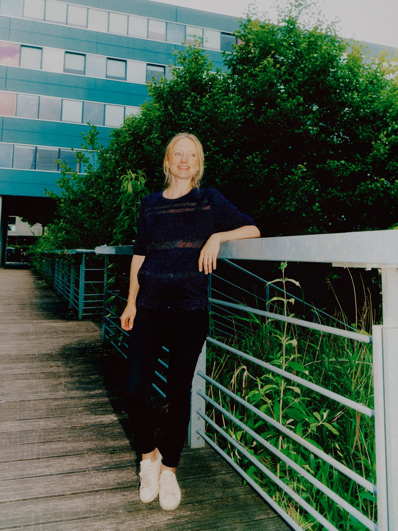 Professional woman with blonde hair and glasses in dark top leaning against metal railing on wooden walkway outdoors
