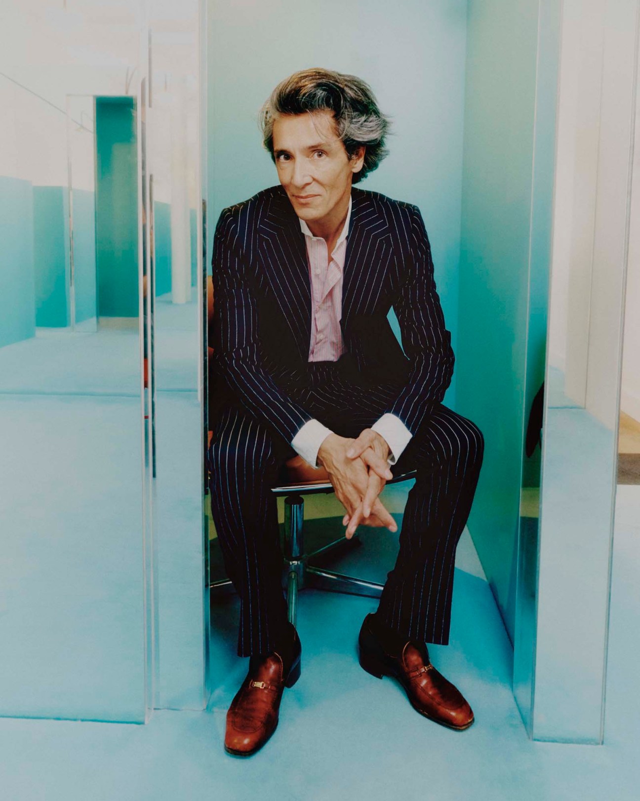 Nicolas Gabard sits in a modern interior wearing a dark pinstripe suit with brown leather shoes