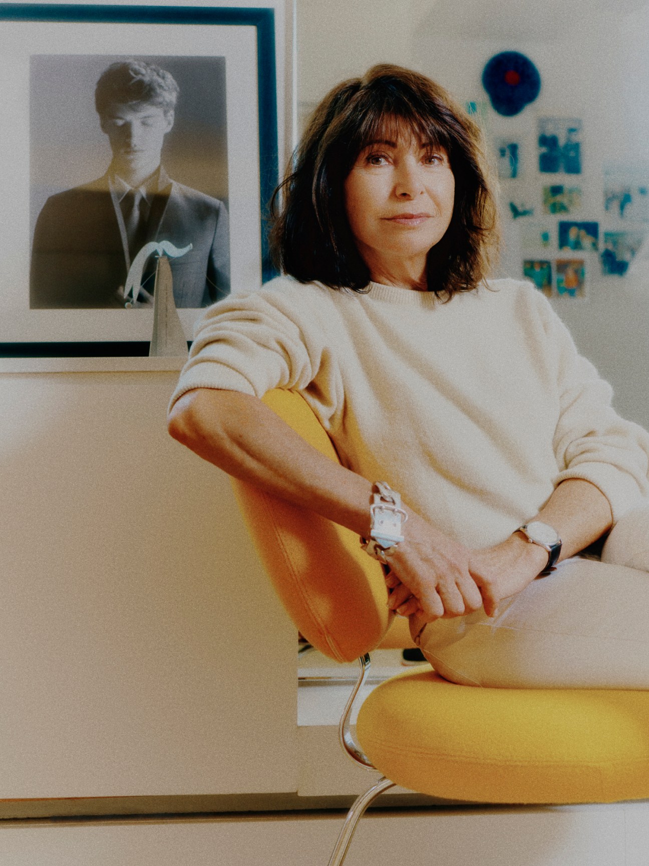 Véronique Nichanian sitting in yellow chair wearing cream sweater in office with framed photos on wall