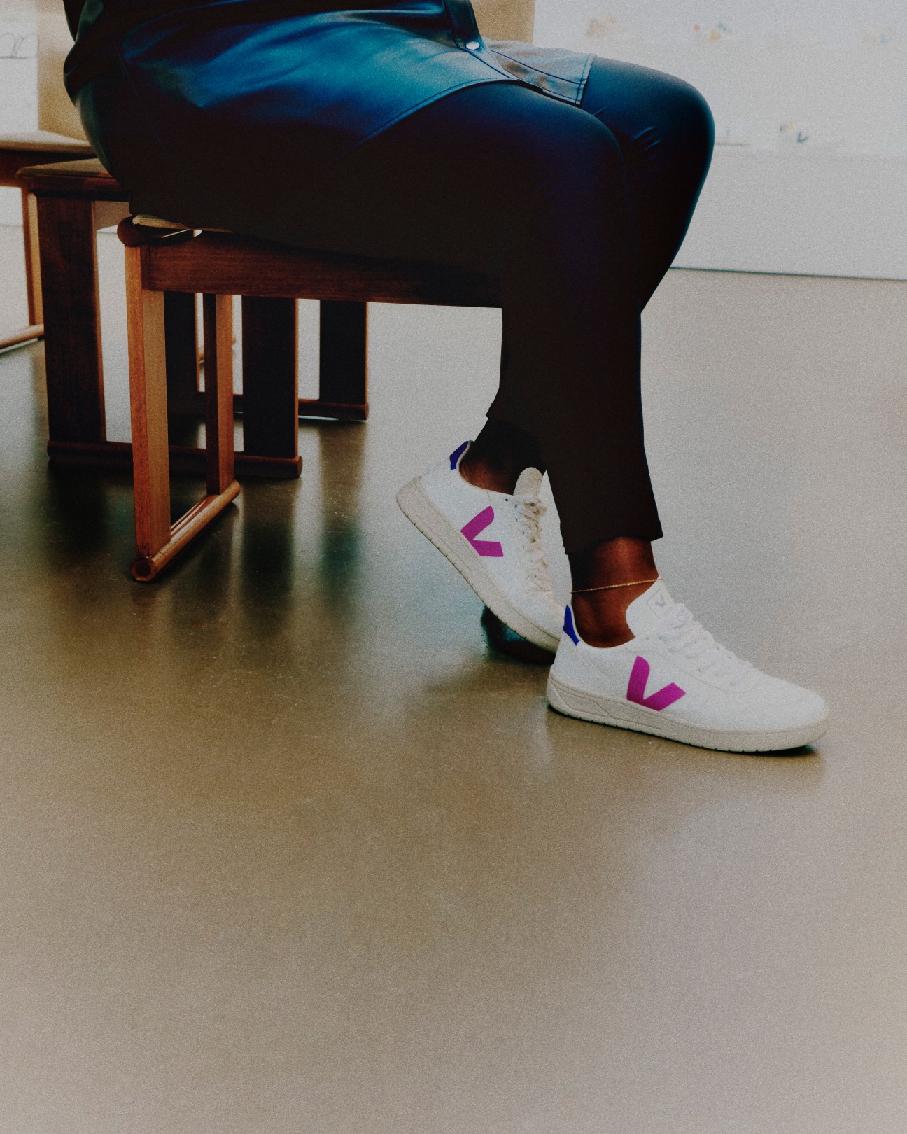Person in dark pants and white Veja sneakers with purple V logo sitting at wooden table