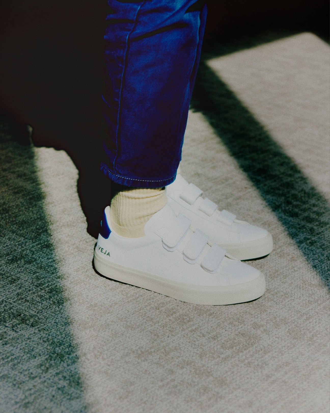 Person wearing white Veja sneakers with velcro straps and blue jeans, standing on striped pavement