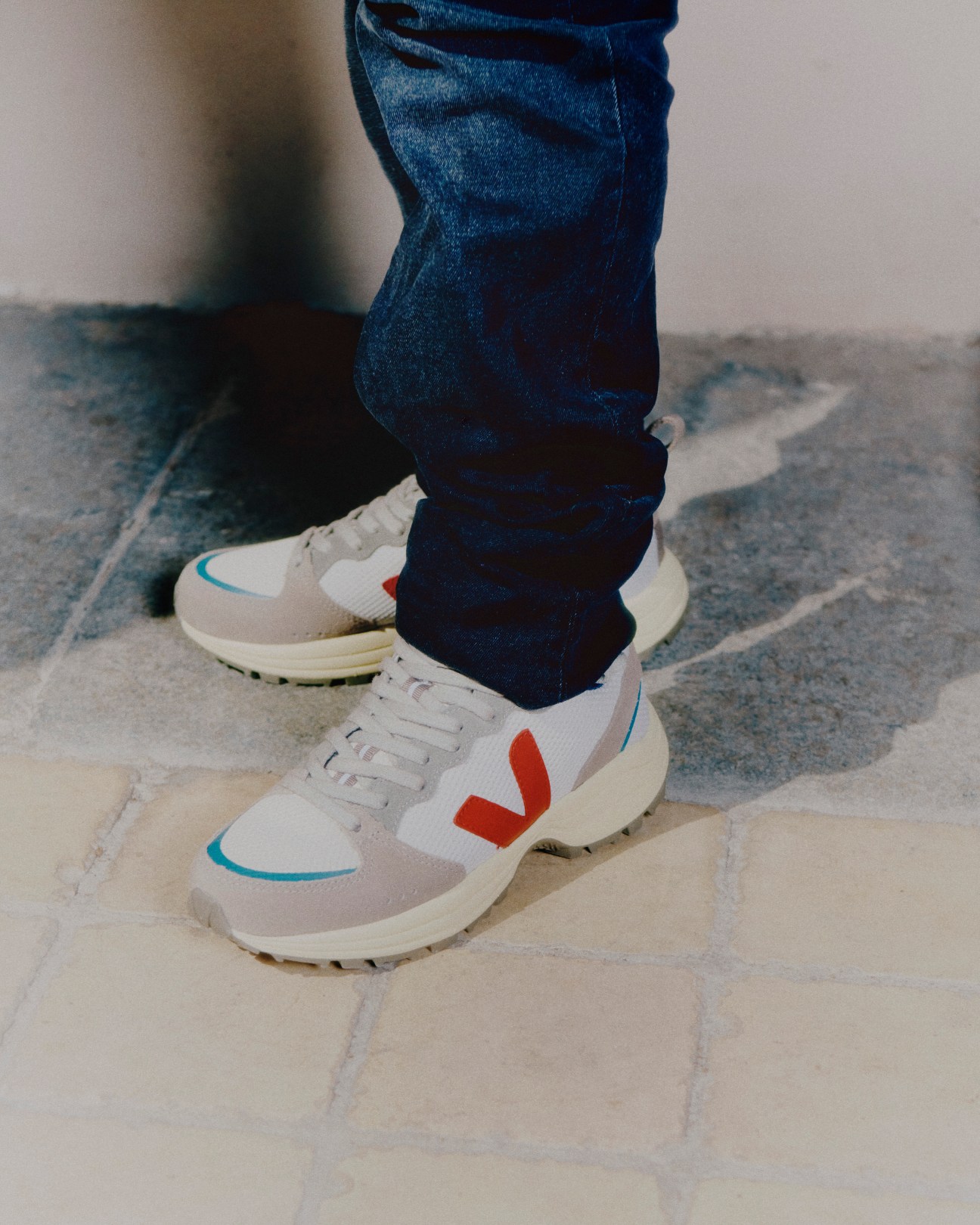 Person wearing white Veja sneakers with red V logo and blue accents, paired with dark jeans on tiled floor