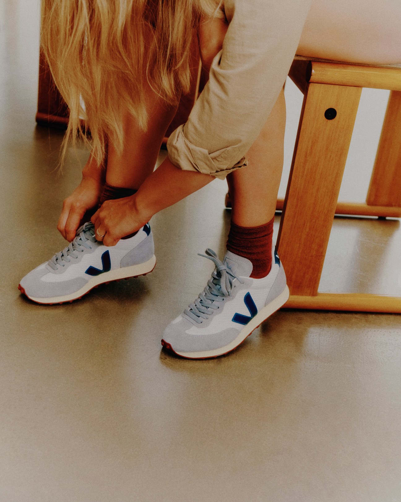 Person in beige top tying laces on white Veja sneakers with blue V logo while sitting on wooden chair