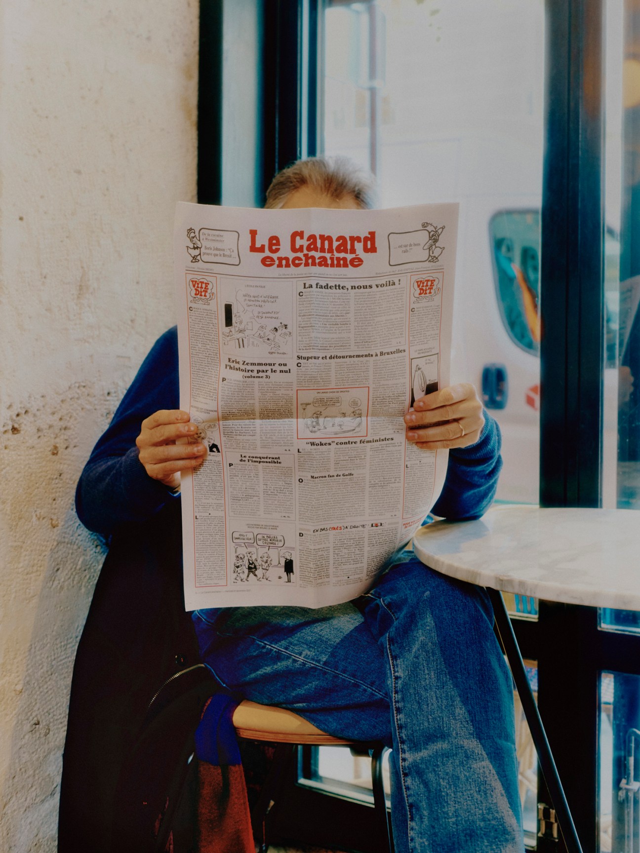 Person reading Le Canard Enchaîné newspaper while sitting at a table by a window