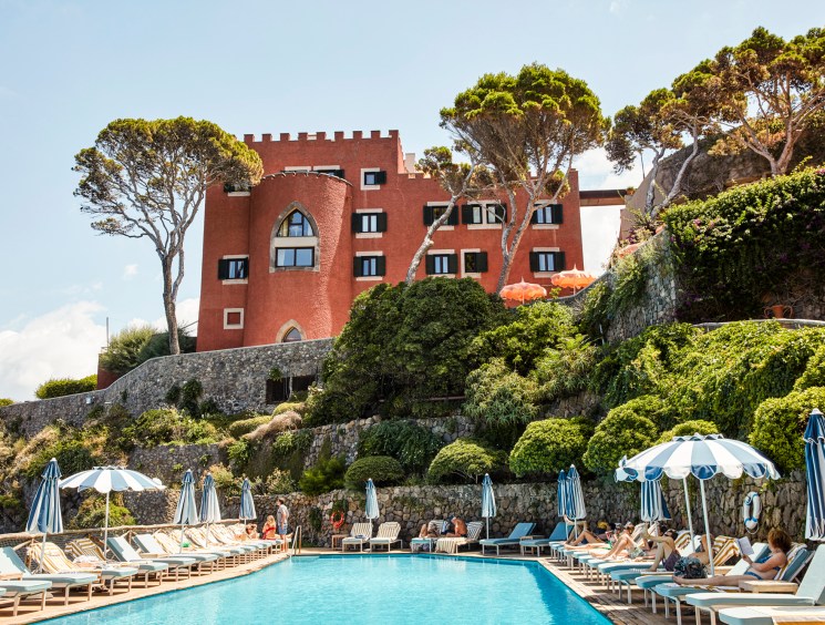 Red Mediterranean hotel building with crenellated towers overlooking a pool area with blue and white umbrellas