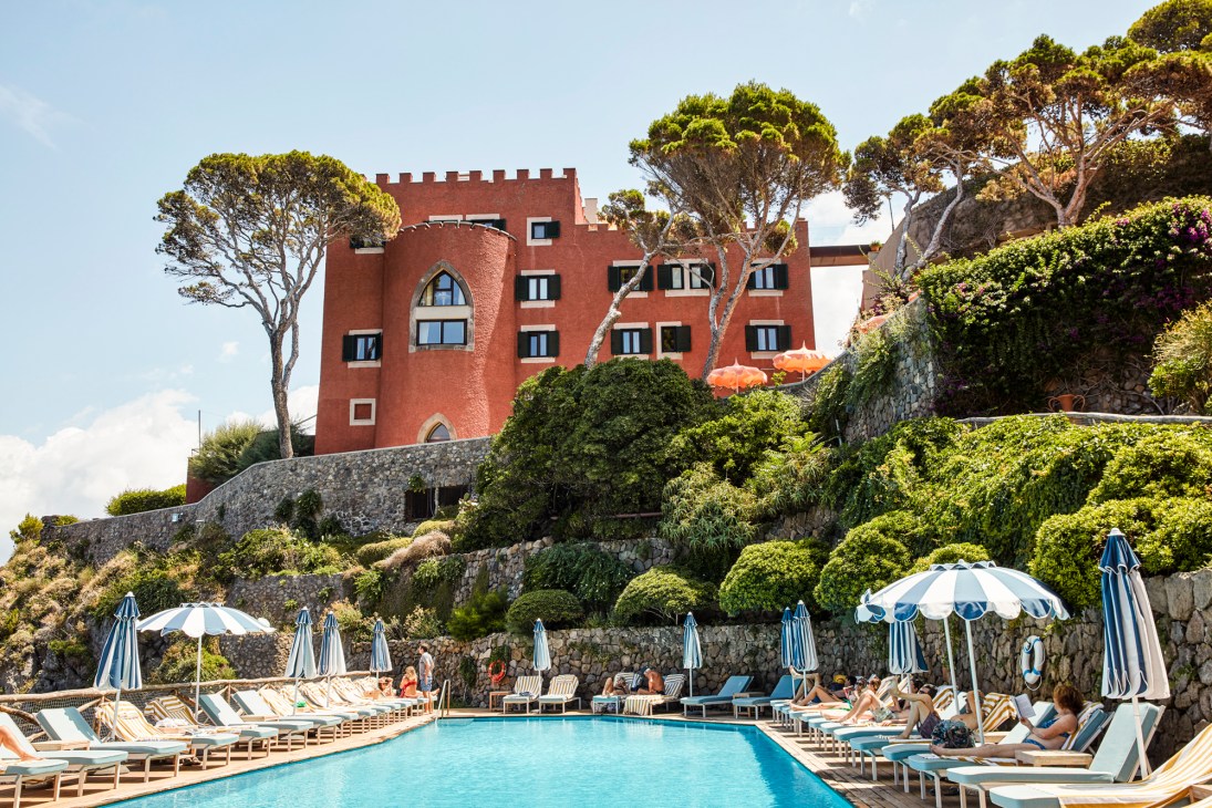 Red Mediterranean hotel building with crenellated towers overlooking a pool area with blue and white umbrellas