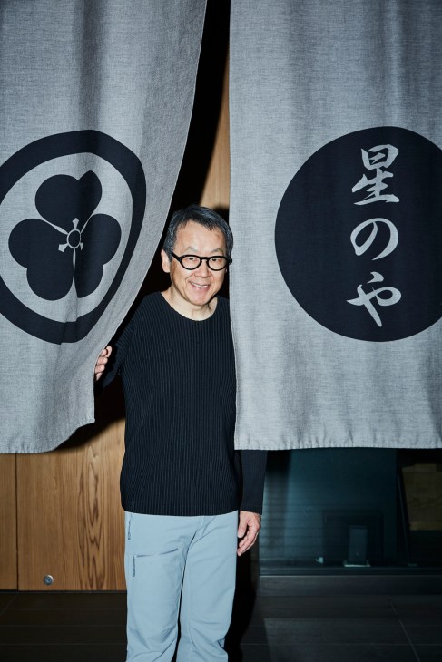 Yoshiharu Hoshino, CEO of Hoshino Resorts, stands between two fabric banners with Japanese characters and crests.