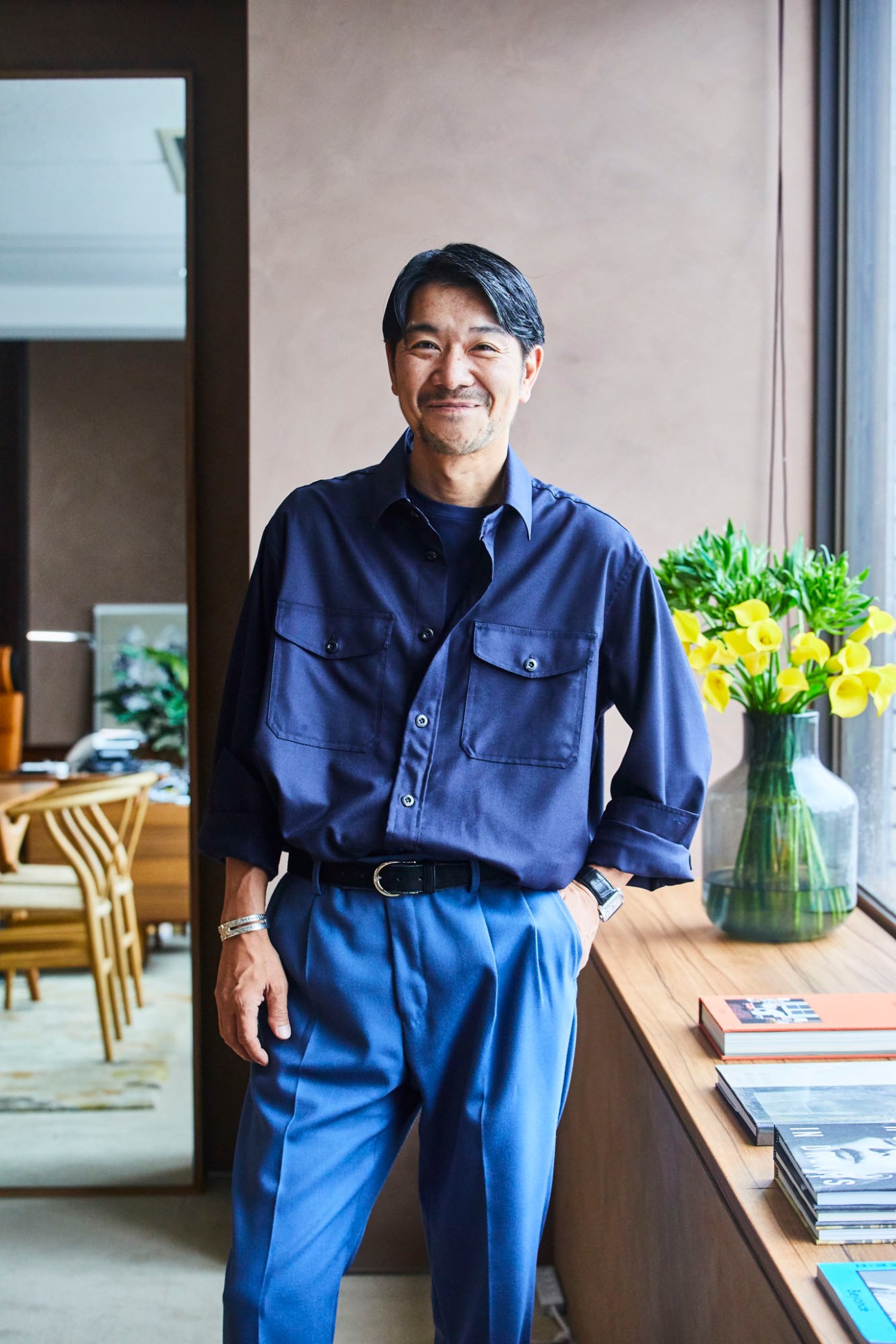 Yoshinori Matsuzaki standing in a modern office wearing a navy blue shirt and pants with yellow flowers on a desk
