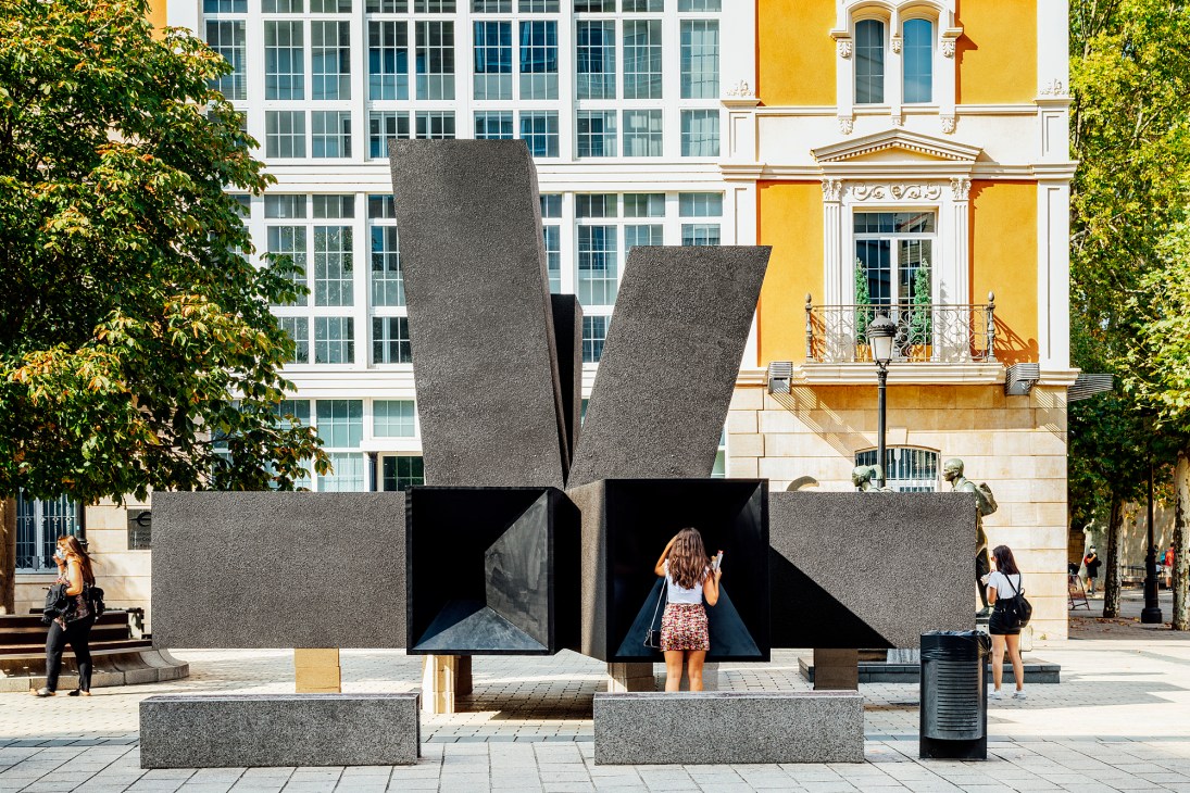 Gray geometric sculptural installation with angular prismatic forms and dark openings in an urban plaza