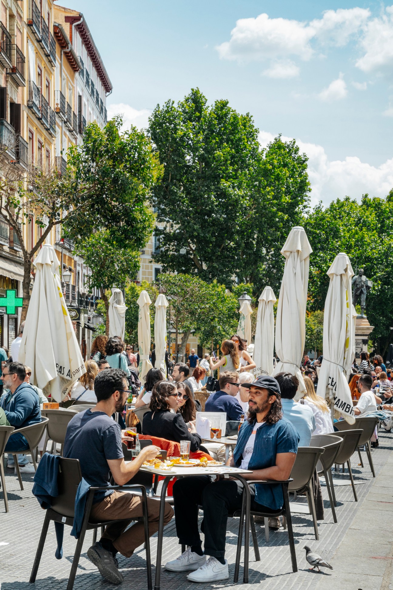 People dining at outdoor restaurant tables with white umbrellas on a sunny plaza in Madrid