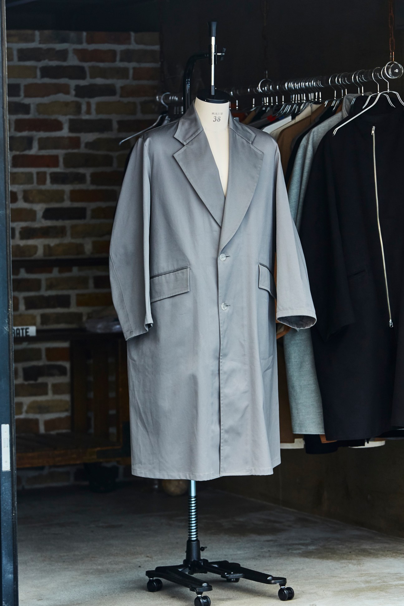 Light grey tailored coat on dress form with other garments on rack behind it.