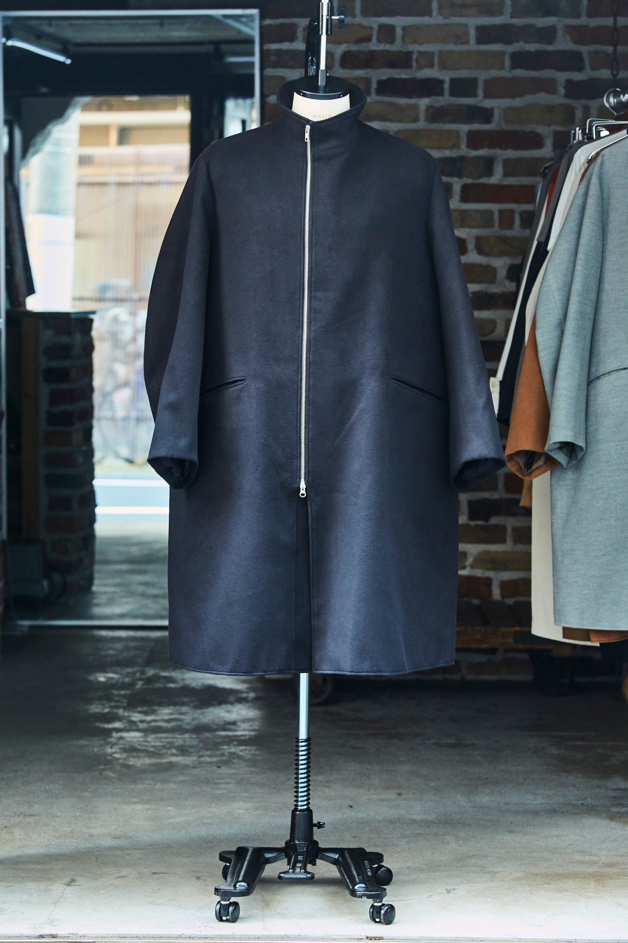 Navy blue zip-up coat displayed on mannequin in brick atelier setting.