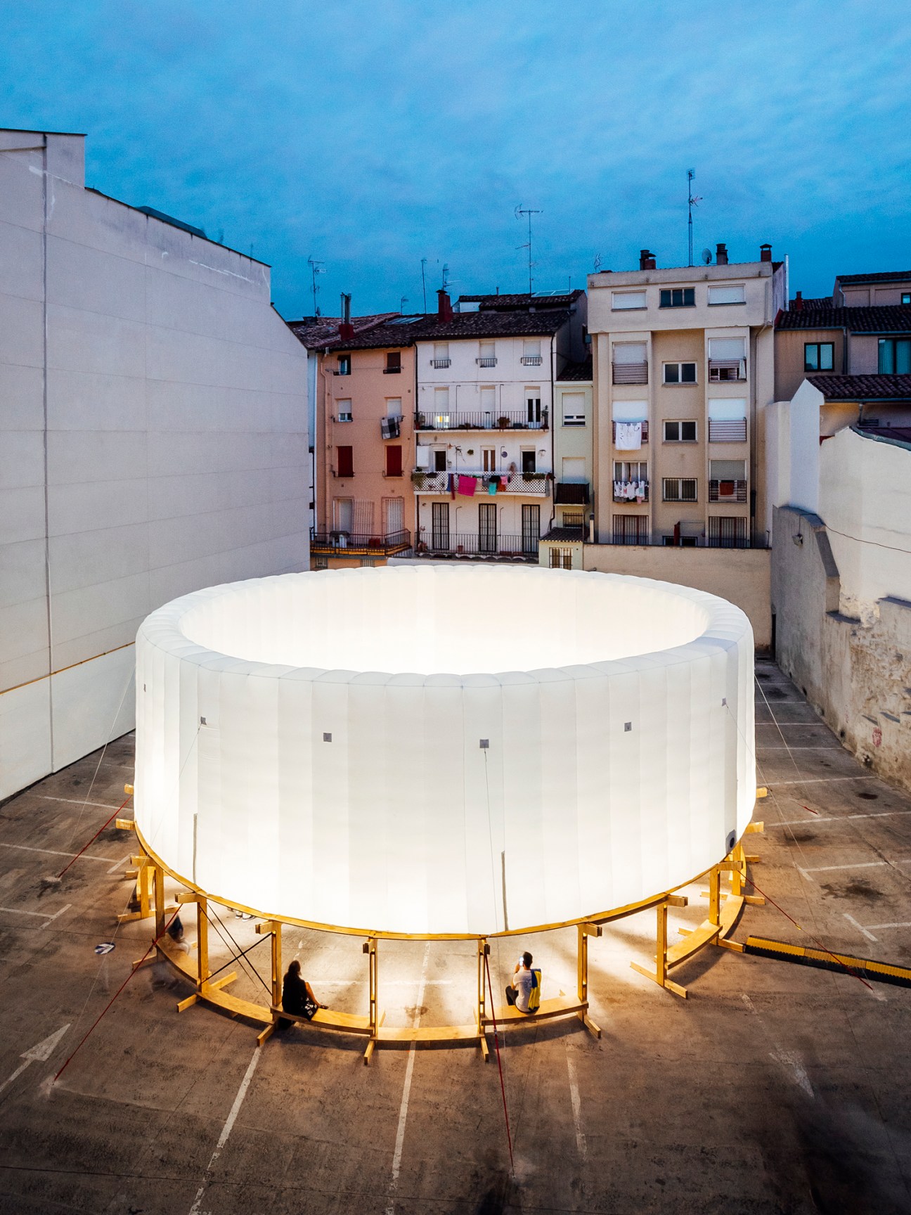 Large white cylindrical installation with illuminated yellow scaffolding base in urban plaza at dusk