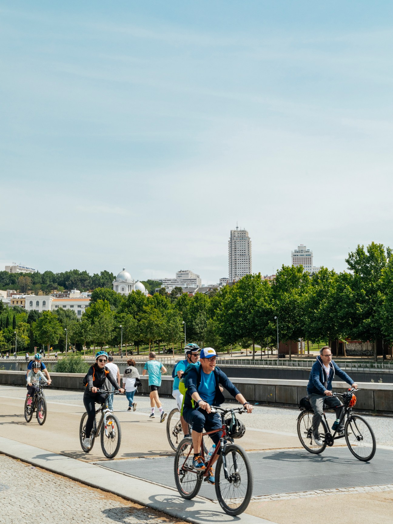 People cycling and walking on a waterfront promenade with green trees and city skyline in the background