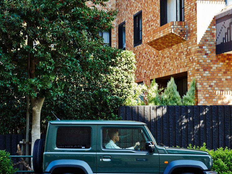 A person driving a vintage green 4WD past a lush tree and modern brick apartment building on a quiet street.