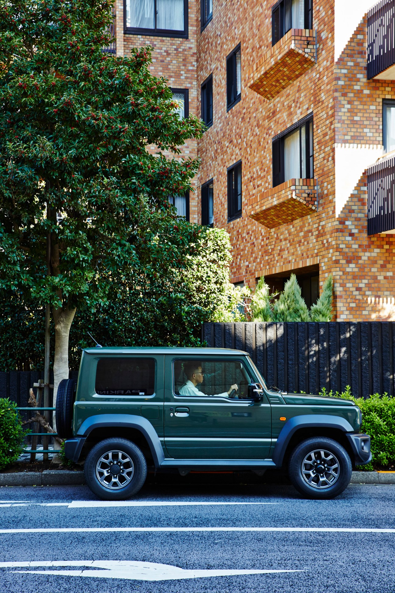 Green Suzuki Jimny SUV parked on street in front of modern brick apartment building with landscaping