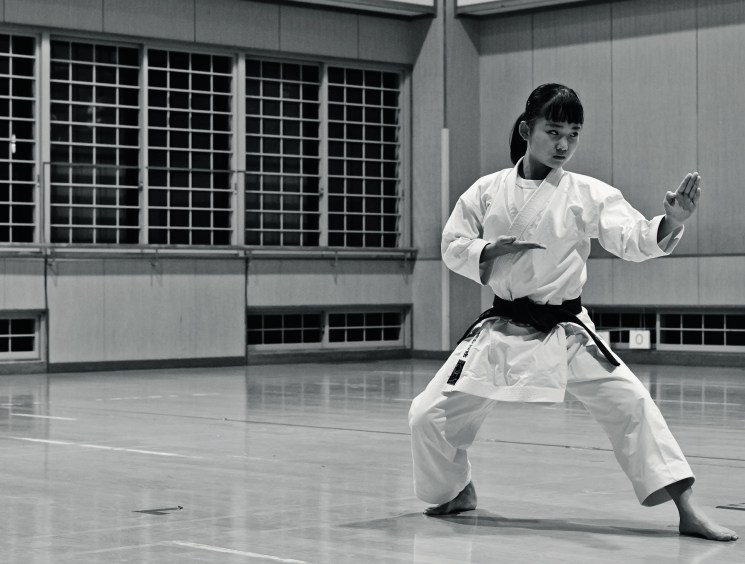Black and white photo of a young woman in a white gi and black belt performing a karate kata in a dojo.