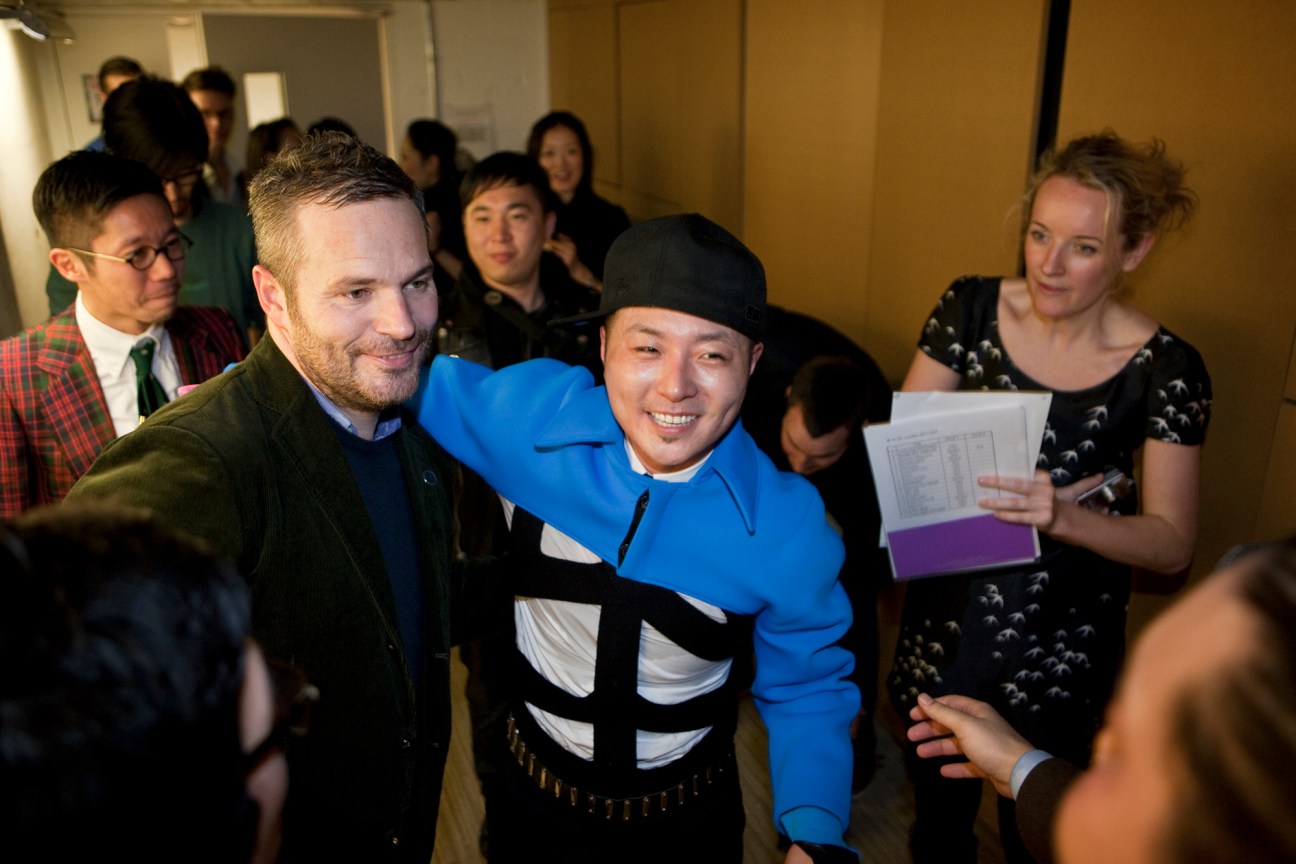 Man in green jacket poses with person in blue and white striped outfit at crowded indoor event