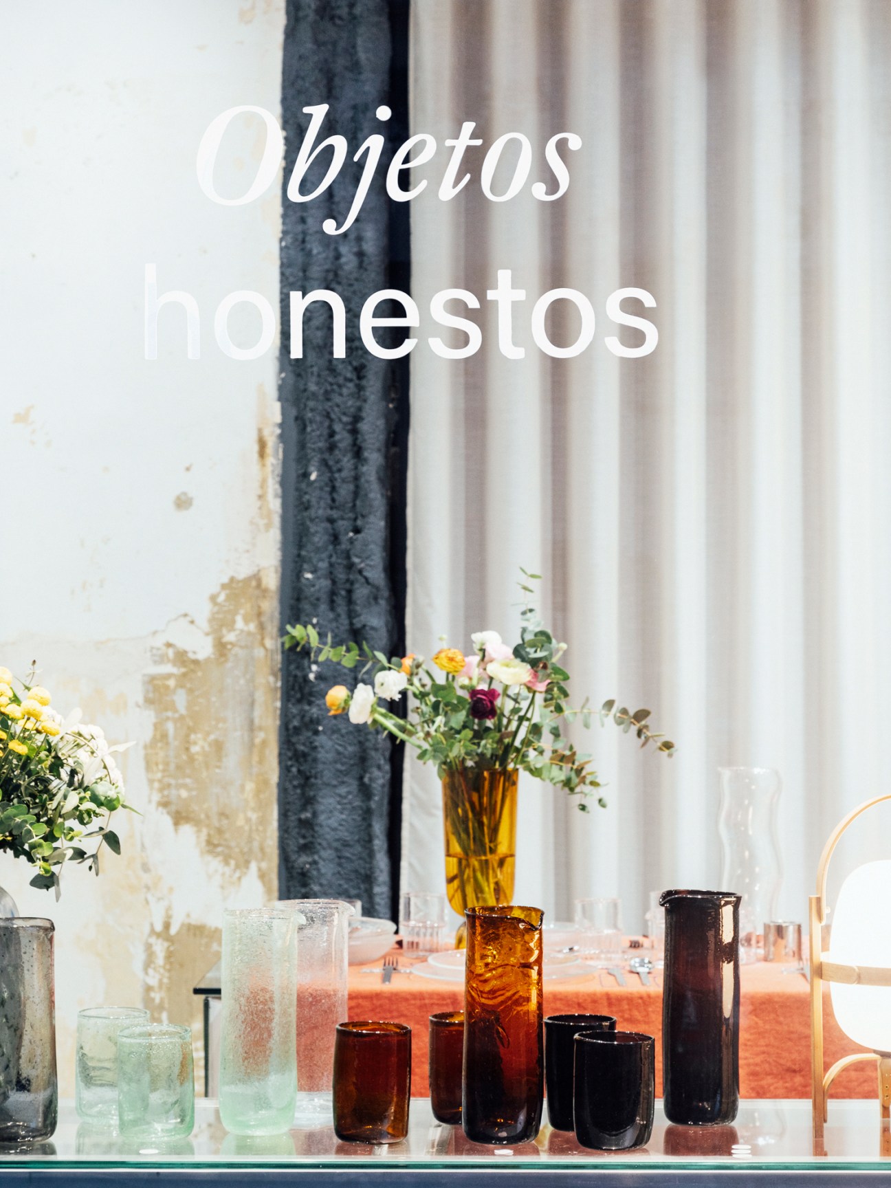 Collection of colorful glassware and ceramic vessels arranged on a glass table near windows