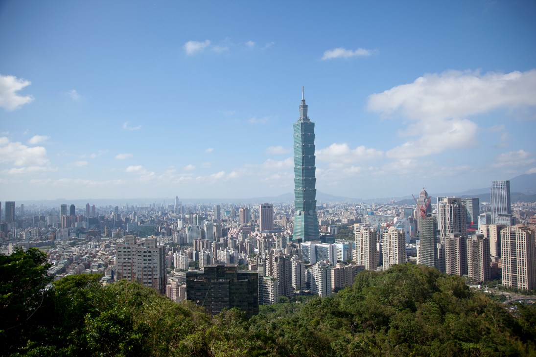 Taipei 101 skyscraper rises above a dense urban skyline of residential and commercial buildings under a blue sky with white clouds.