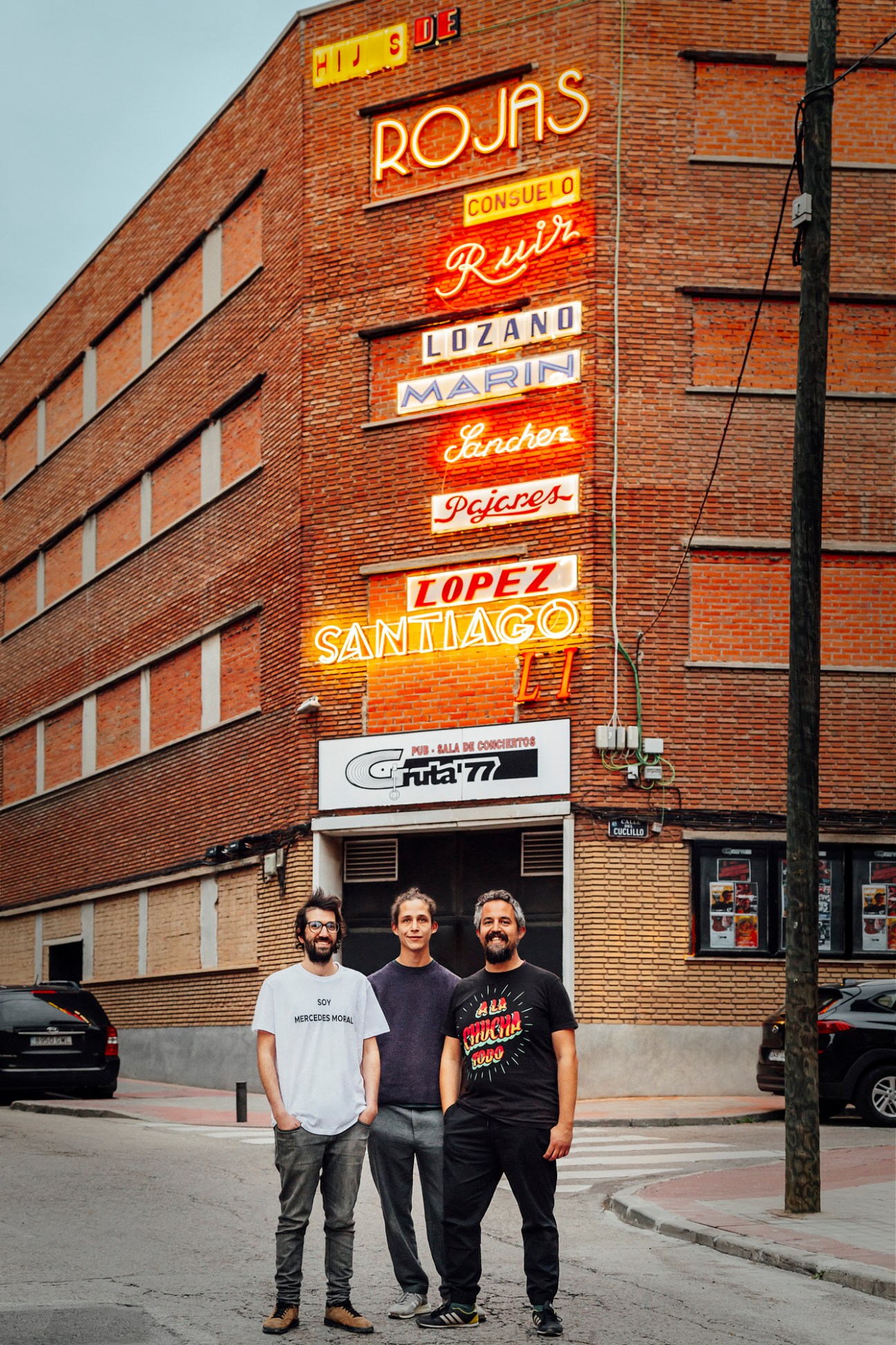 Three men standing in front of brick building with vintage neon signs including Rojas, Consuelo, Ruiz, Lozano, Marin