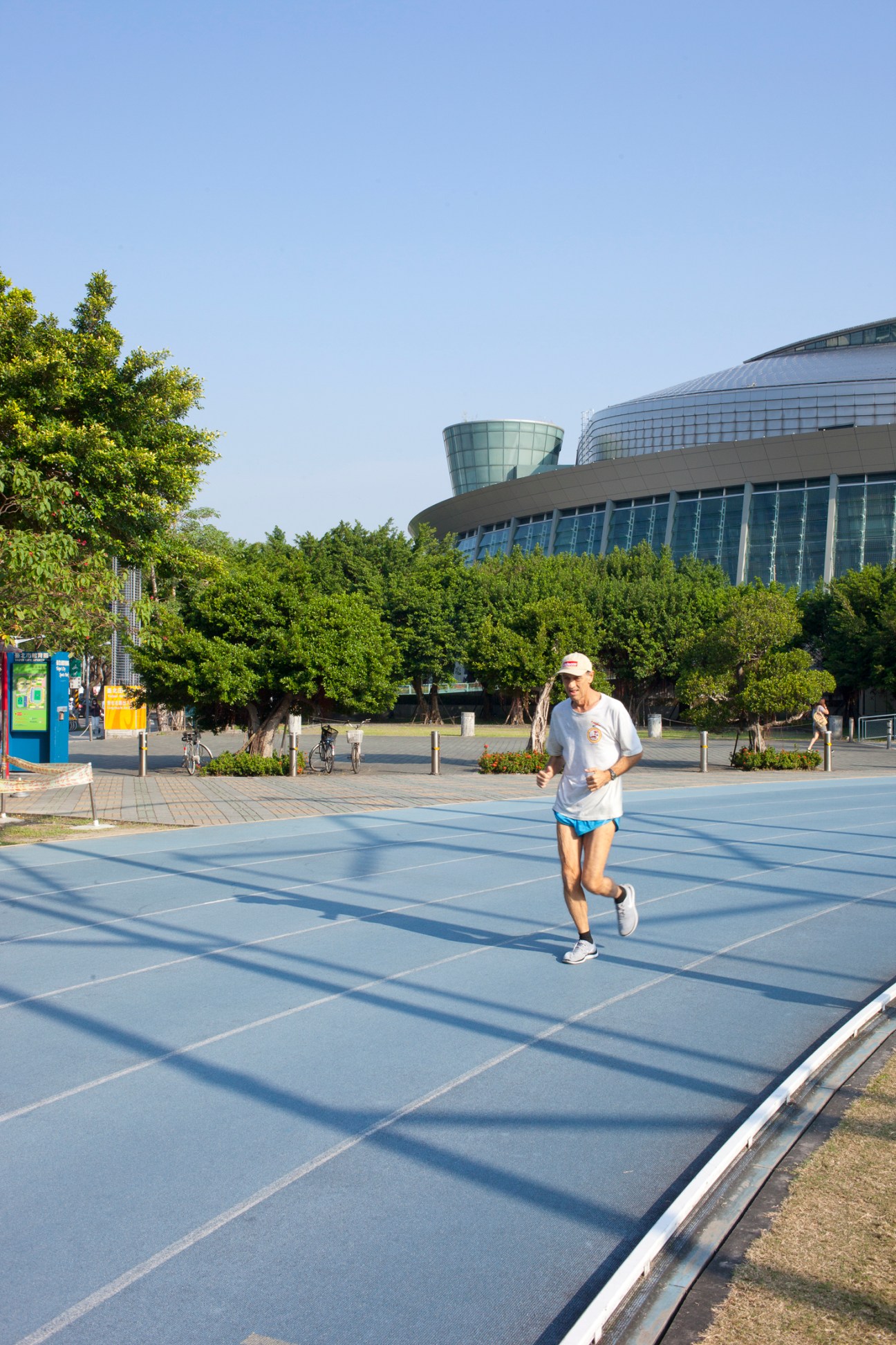 Man jogging on blue track at Taipei Sports Arena with modern curved building in background