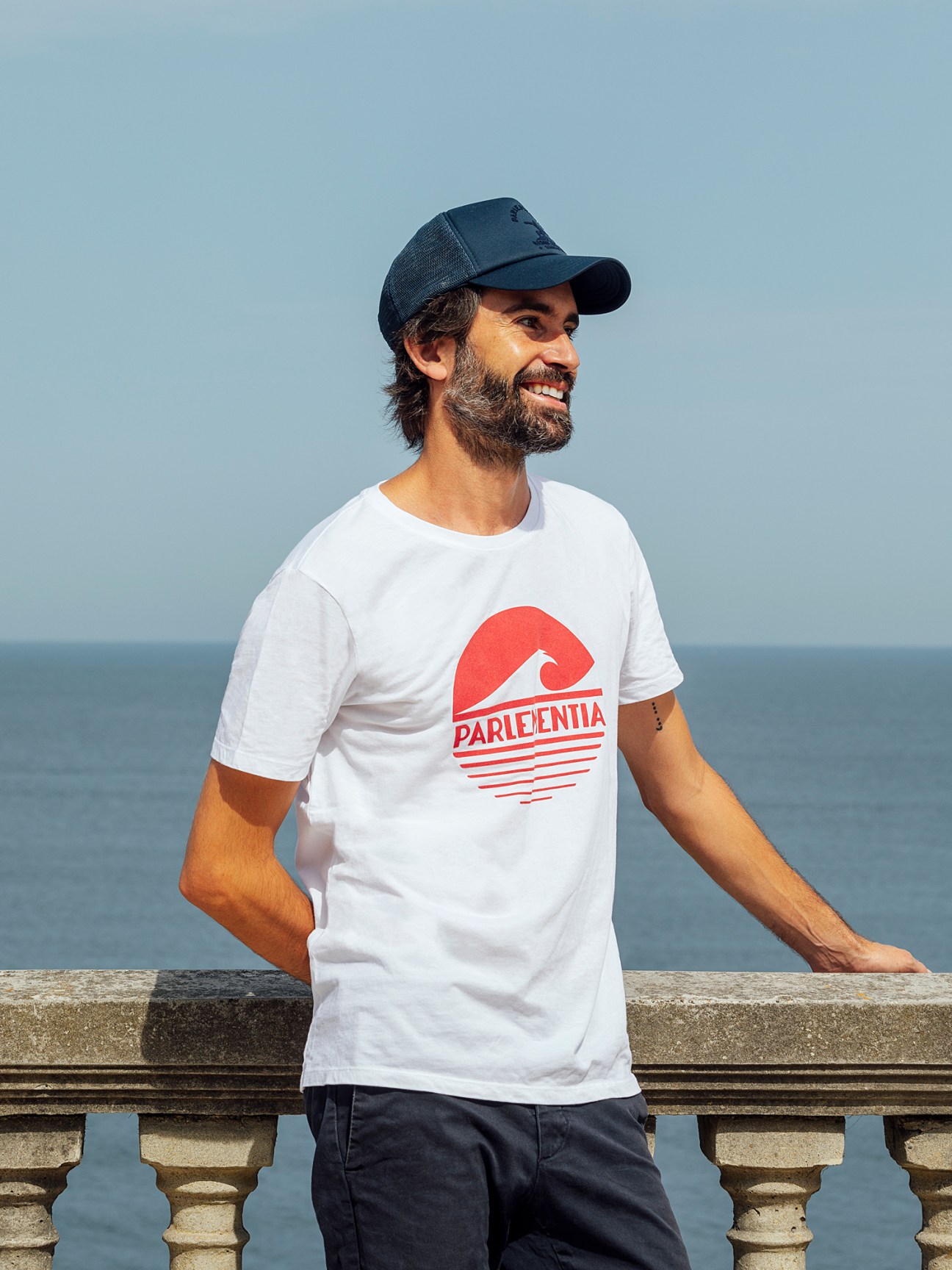 Man wearing Parlementia white t-shirt and navy cap standing by the sea in Guéthary, France.