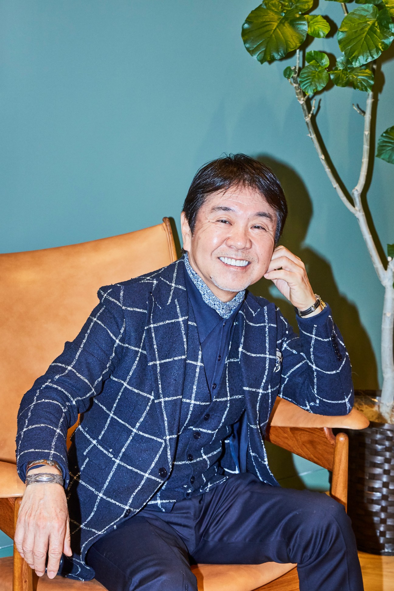 Yo Shitara sitting in a wooden chair wearing a navy checkered shirt, smiling at the camera