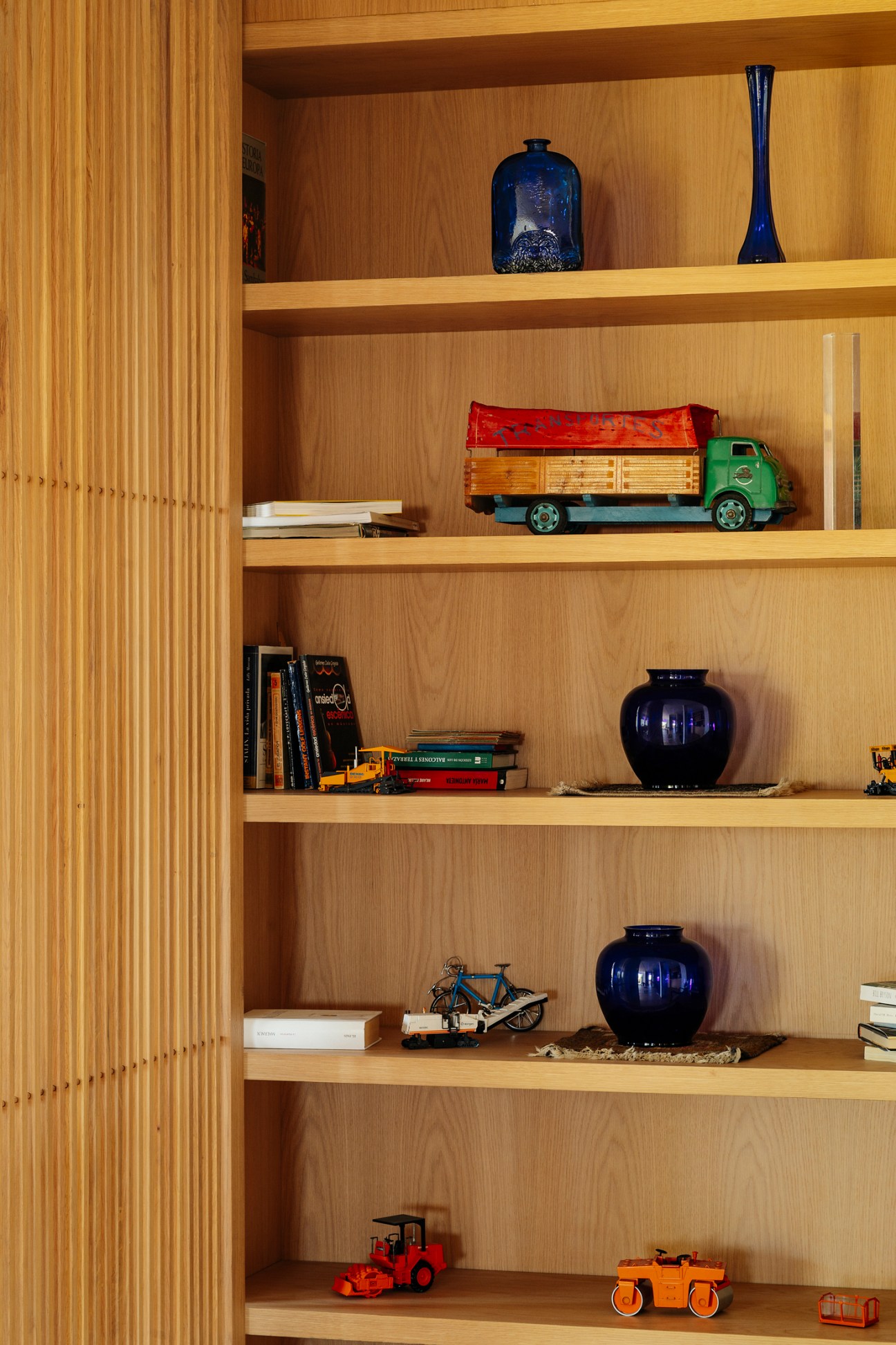 Wooden shelving unit displaying blue ceramic vessels, toy vehicles, and books in organized arrangement.