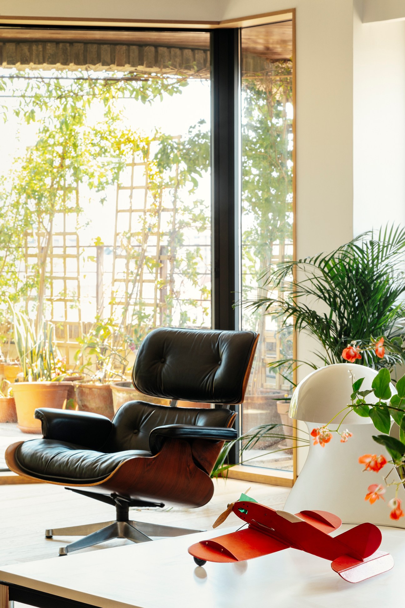 Modern living room interior with black leather Eames chair, glass table, and large windows overlooking greenery