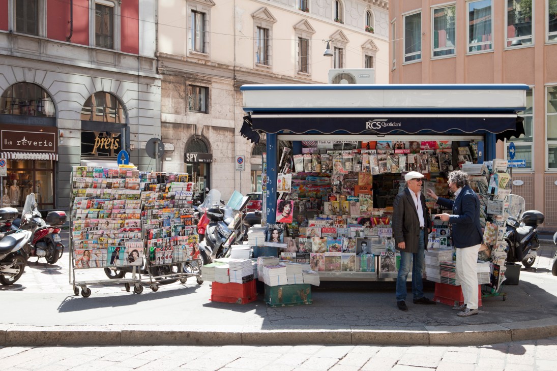 A busy Italian newsstand with magazines and newspapers displayed, two men conversing beside the blue kiosk